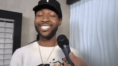 A Man Wearing A Hat Is Holding A Microphone In His Hand And Smiling . GIF
