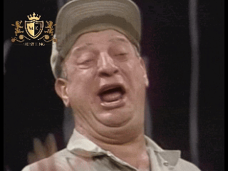 A Man Wearing A Hat Is Making A Funny Face In Front Of A Shield With The Letter K On It GIF