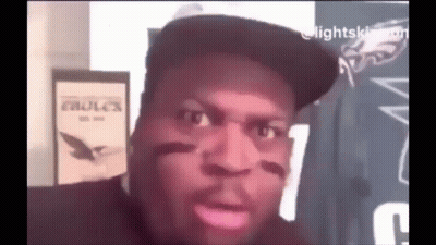 A Man Wearing A Hat Is Making A Funny Face While Sitting In Front Of A Sign That Says Eagles . GIF