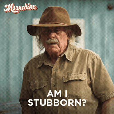 A Man Wearing A Hat Says " Am I Stubborn " In Front Of A Blue Wall GIF