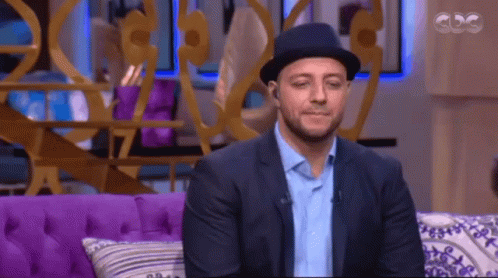 A Man Wearing A Hat Sits On A Purple Couch In Front Of A Cdg Sign GIF