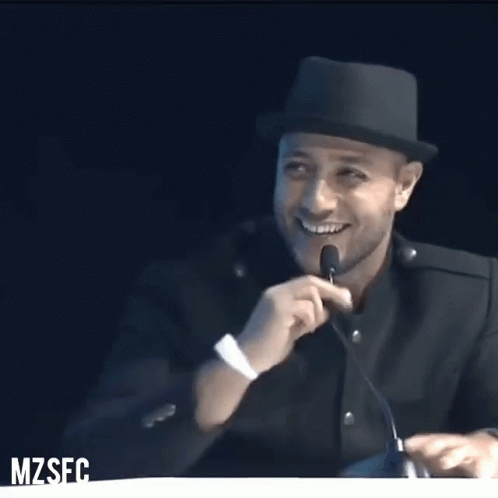 A Man Wearing A Hat Speaking Into A Microphone With The Word Mzses On The Bottom Right GIF