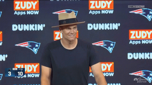 A Man Wearing A Hat Stands In Front Of A Dunkin ' Advertisement GIF
