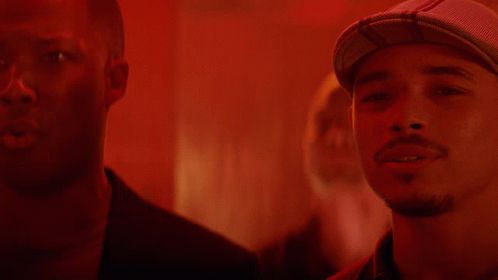 A Man Wearing A Hat Stands Next To Another Man In A Red Room GIF