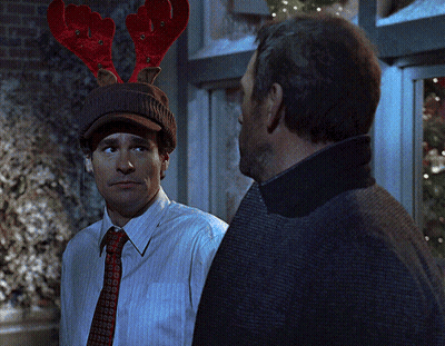 A Man Wearing A Hat With Reindeer Antlers Looks At Another Man GIF