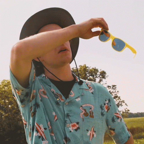 A Man Wearing A Hawaiian Shirt And A Hat Holds A Pair Of Yellow Sunglasses GIF