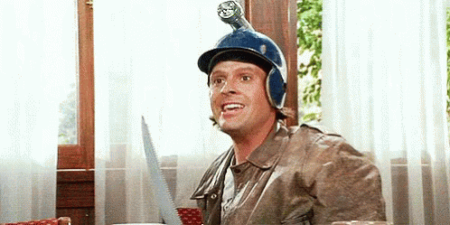 A Man Wearing A Helmet And A Jacket Is Sitting At A Table With A Saw On His Head . GIF