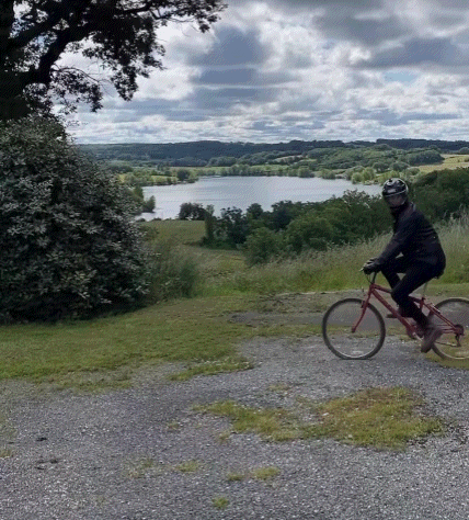 A Man Wearing A Helmet Is Riding A Bike On A Gravel Road With A Lake In The Background GIF
