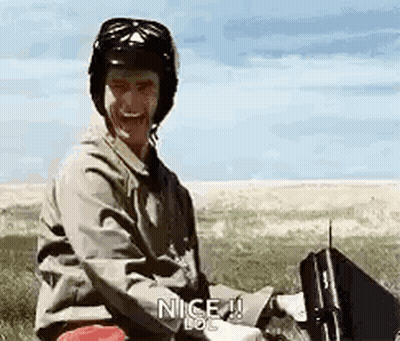 A Man Wearing A Helmet Is Sitting In A Field With A Briefcase And A Gun . GIF