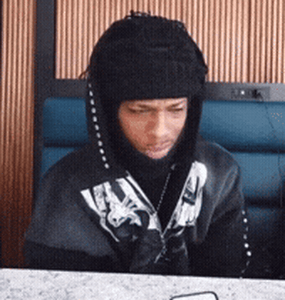 A Man Wearing A Hooded Jacket And A Beanie Is Sitting At A Table . GIF