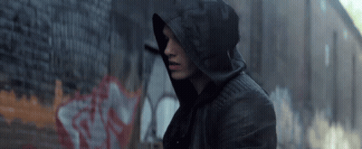 A Man Wearing A Hooded Jacket Is Standing In Front Of A Brick Wall With Graffiti On It GIF