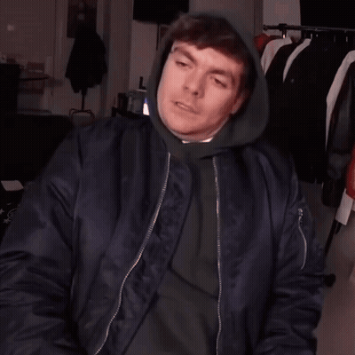 A Man Wearing A Hoodie And A Bomber Jacket Is Making A Hand Gesture GIF