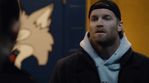 A Man Wearing A Hoodie And A Hat Stands In Front Of A Wolf Emblem GIF