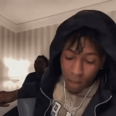 A Man Wearing A Hoodie And A Necklace Is Sitting In A Room With His Eyes Closed . GIF