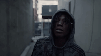 A Man Wearing A Hoodie Is Standing In A Dark Alleyway . GIF