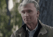 A Man Wearing A Jacket And A Plaid Shirt Is Standing In Front Of A Tree . GIF
