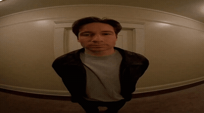A Man Wearing A Leather Jacket And A Grey Shirt Stands In A Hallway GIF