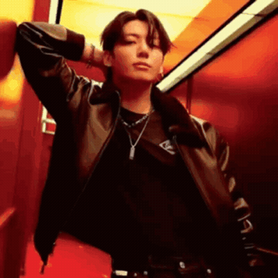 A Man Wearing A Leather Jacket And A Necklace Is Leaning Against A Red Wall . GIF