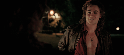 A Man Wearing A Leather Jacket And A Necklace Smiles At Another Man GIF