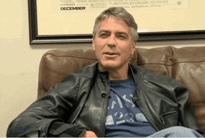 A Man Wearing A Leather Jacket Sits On A Couch In Front Of A Framed Picture That Says December GIF