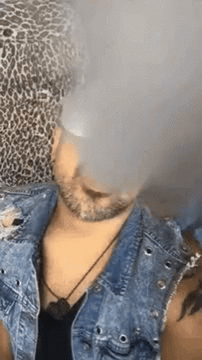 A Man Wearing A Leopard Print Hat And A Denim Jacket Is Smoking An Electronic Cigarette . GIF