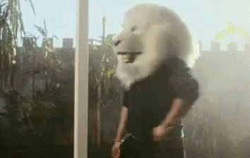 A Man Wearing A Lion Mask Is Standing In Front Of A Window And Smoking A Cigarette . GIF