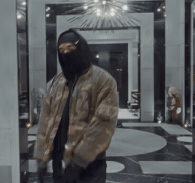 A Man Wearing A Mask And A Bomber Jacket Is Standing In A Room . GIF