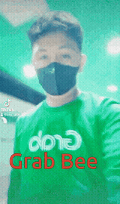 A Man Wearing A Mask And A Green Shirt That Says Grab Bee Is Taking A Selfie . GIF