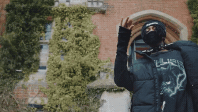 A Man Wearing A Mask And A Supreme Hat Stands In Front Of A Building GIF