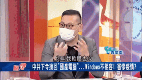 A Man Wearing A Mask And Glasses Is Sitting At A Table With Chinese Writing On The Screen Behind Him GIF
