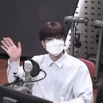A Man Wearing A Mask And Glasses Is Sitting In Front Of A Microphone And Waving . GIF