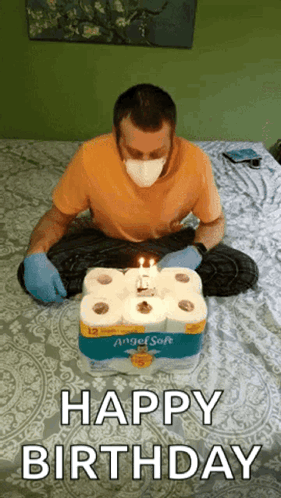 A Man Wearing A Mask And Gloves Is Sitting On A Bed With A Birthday Cake Made Out Of Toilet Paper Rolls . GIF