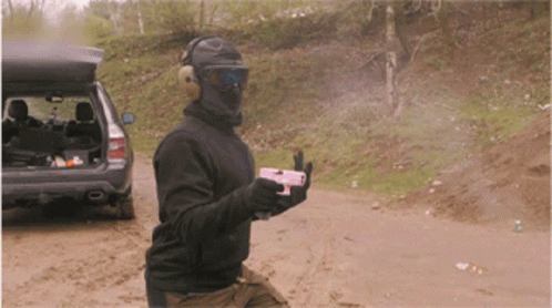 A Man Wearing A Mask And Goggles Is Holding A Gun In Front Of A Car GIF