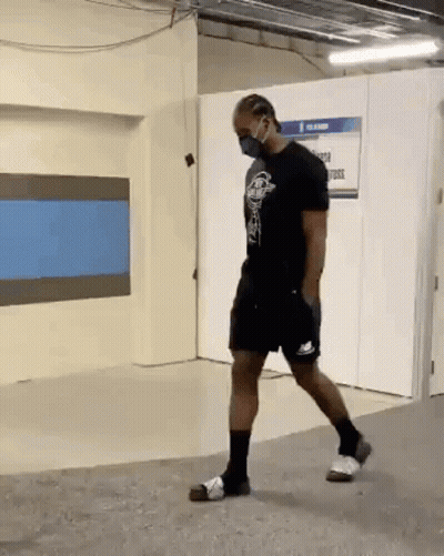 A Man Wearing A Mask And Shorts Is Walking In A Room . GIF