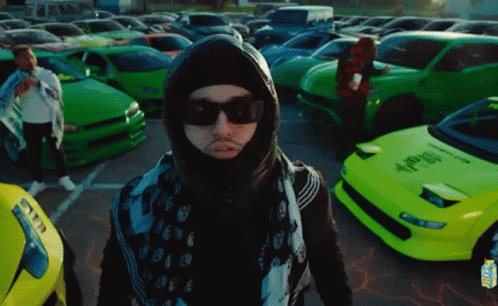 A Man Wearing A Mask And Sunglasses Is Standing In Front Of A Parking Lot Full Of Green Cars GIF