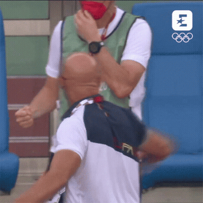 A Man Wearing A Mask Is Being Helped By Another Man With The Olympic Logo In The Background GIF