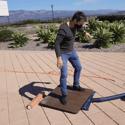 A Man Wearing A Mask Stands On A Wooden Table Outside GIF