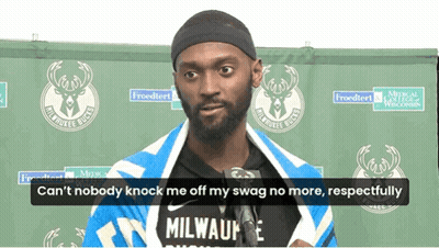 A Man Wearing A Milwaukee Bucks Shirt Is Standing In Front Of A Green Backdrop GIF