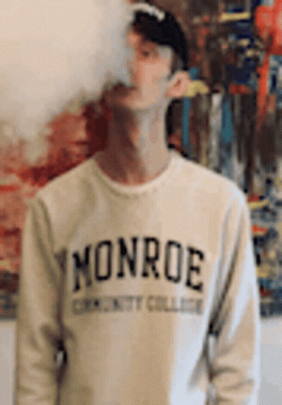 A Man Wearing A Monroe Sweatshirt Is Smoking A Cigarette In Front Of A Painting . GIF