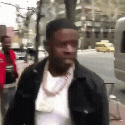 A Man Wearing A Necklace And A Jacket Is Walking Down A Street . GIF