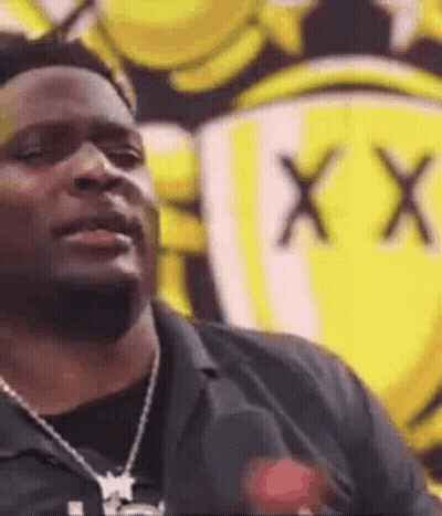 A Man Wearing A Necklace Is Standing In Front Of A Yellow Sign . GIF