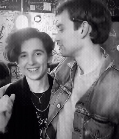A Man Wearing A Necklace That Says ' The Singer ' On It Stands Next To Another Man GIF