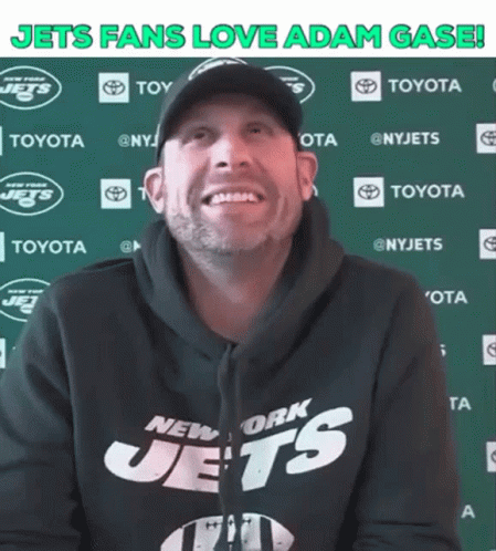 A Man Wearing A New York Jets Sweatshirt Smiles In Front Of A Green Wall GIF