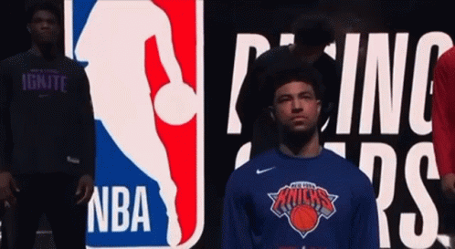A Man Wearing A New York Knicks Shirt Is Standing In Front Of A Nba Logo GIF