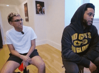 A Man Wearing A No Cys Hoodie Sits Next To A Man Wearing Shorts GIF
