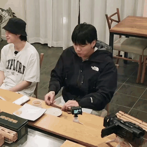 A Man Wearing A North Face Jacket Sits At A Table With Another Man GIF