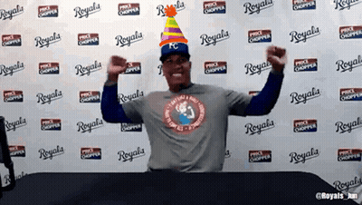 A Man Wearing A Party Hat Is Standing In Front Of A Wall That Says Royals On It GIF