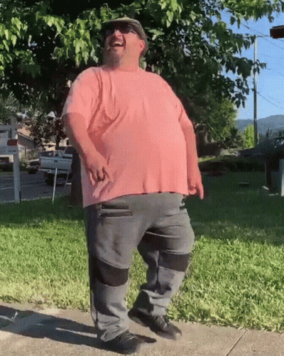 A Man Wearing A Pink Shirt And Grey Pants Is Standing On A Sidewalk GIF