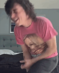 A Man Wearing A Pink Shirt With A Picture Of A Woman On It Is Sitting On A Bed GIF