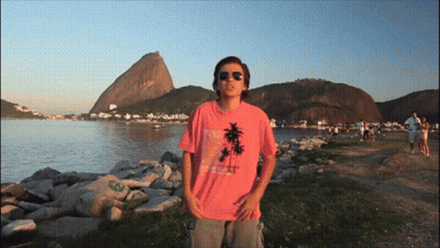 A Man Wearing A Pink Shirt With Palm Trees On It Stands In Front Of A Body Of Water GIF